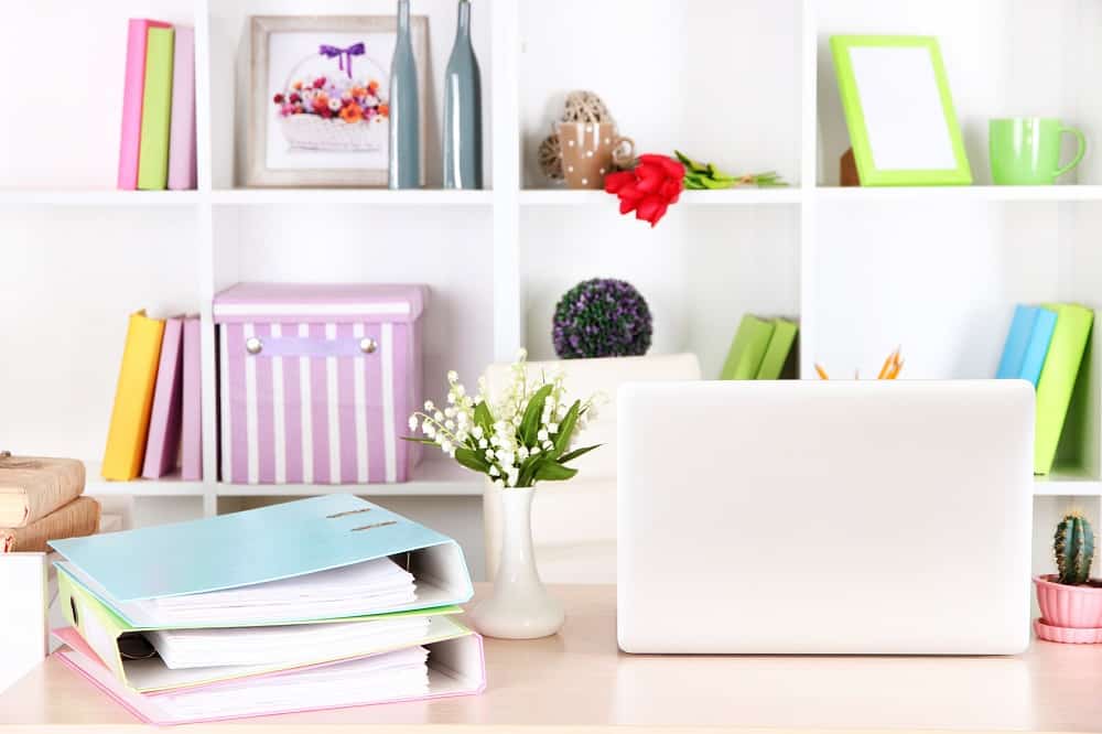 Home Business Ideas for Women. A desk with a laptop and lever-arch files. A white bookcase in the background with books and ornaments on it.