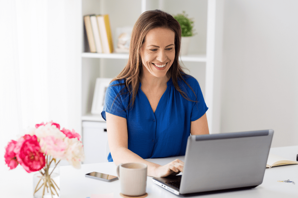 How to Become a Blogger. Woman sitting at a desk with her laptop, phone, mug of coffee and some pink & white flowers.